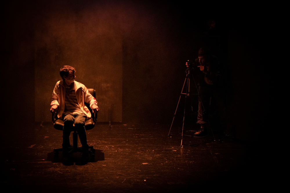 Un comédien est ligoté à une chaise et un autre comédien le filme dans une douche lumineuse orangée, création lumière de Jérôme Baudouin, créateur lumière en danse, théâtre et spectacle vivant